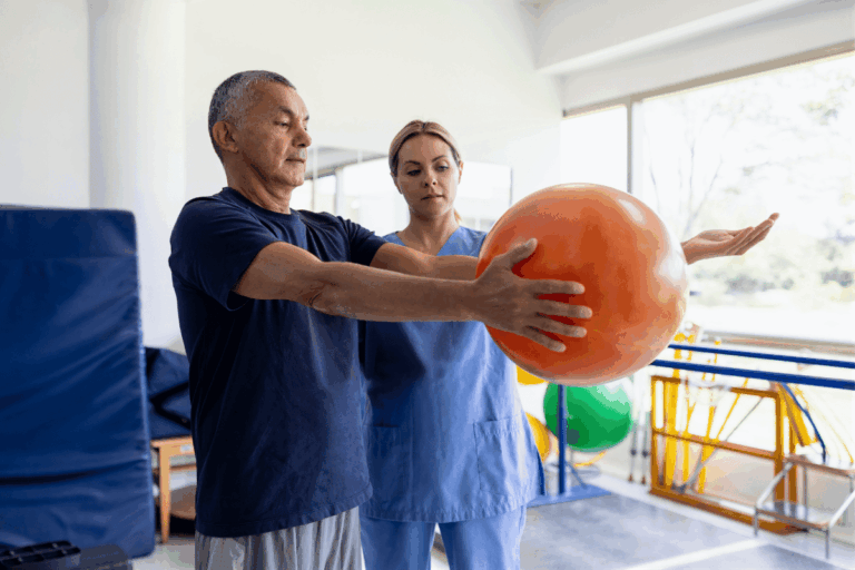Mader MedX Physical therapy assistant guiding an older man through an exercise with an orange therapy ball in a rehabilitation clinic.